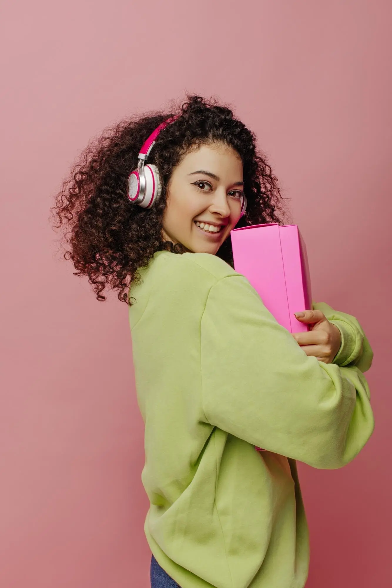 Eine attraktive, hellhäutige junge Frau steht seitlich vor pinkem Hintergrund und hält eine Schachtel an sich. Die Brünette mit Kopfhörern und Jacke lächelt in die Kamera – Emotionenkonzept, Gesichtsausdruck.